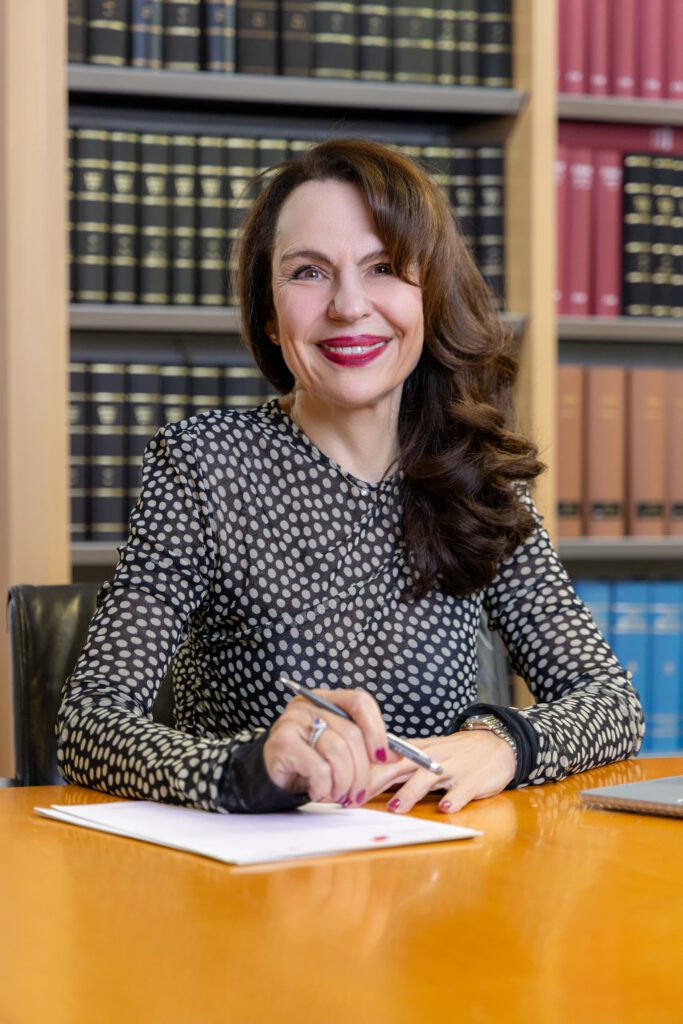 Anne Patsch sitzt am Schreibtisch und unterschreibt ein Dokument, im Hintergrund juristische Fachliteratur